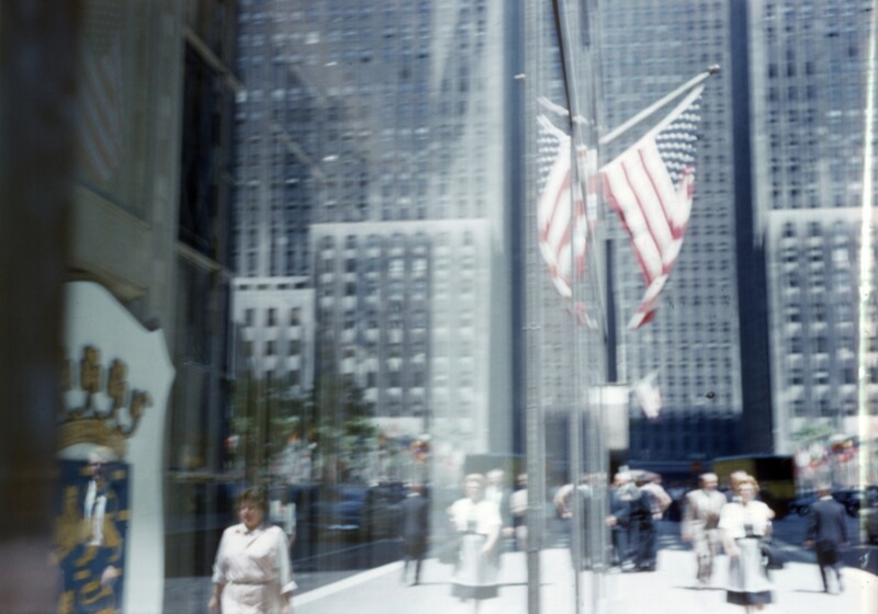A photo of the record -- American Flag in a city with people walking along a building and their reflection in the building windows,  1985